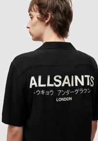 Black short-sleeve shirt with a camp collar featuring bold white text on the back: "ALLSAINTS" and other characters, smooth texture.