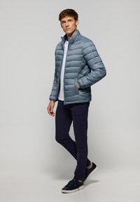 Ljusblå vadderad jacka med hög krage, dragkedjestängning och två sidofickor, bärs över en vit skjorta och marinblå byxor med sneakers.