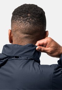 Veste imperméable bleu foncé avec un col haut, montrant une personne ajustant le col. Le tissu semble lisse et léger.