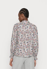 Blusa de manga longa floral com costas franzidas, apresentando uma mistura de pequenos padrões em vermelho, azul e verde sobre um fundo claro. Tecido texturizado.