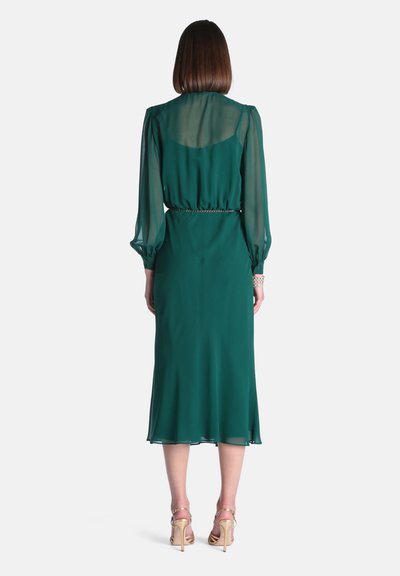 Femme aux cheveux mi-longs portant une robe midi vert émeraude à manches longues avec un dos supérieur transparent et des sandales dorées à talons hauts.