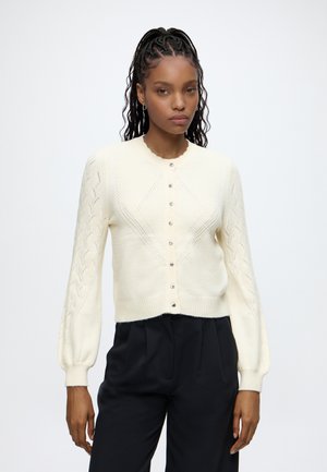 Femme aux cheveux tressés portant un cardigan en tricot crème boutonné avec des manches à motifs et un pantalon noir taille haute sur un fond uni.