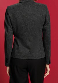 Kvinna som bär en åtsittande mörkgrå texturerad kavaj och svarta byxor, stående mot en enfärgad röd bakgrund, sedd bakifrån.