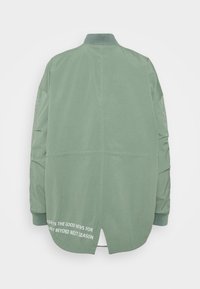 Przesadnie duża, jasnozielona kurtka z długimi rękawami, prążkowanym kołnierzem i mankietami oraz białym napisem blisko dolnego szwu z tyłu.