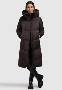 Dolga črna puhovka s sijočo površino, oversize kapuco, pritrdilne gumbe, zadrgo in prešito površino; v kombinaciji s črnimi škornji do kolen.