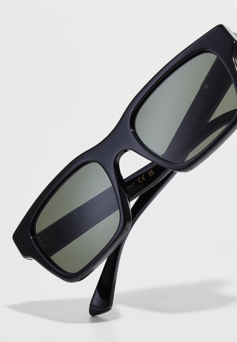 Black oversized sunglasses with green tinted lenses, rectangular shape, thick frames, featuring a sleek finish and logo detailing on the inner arm.
