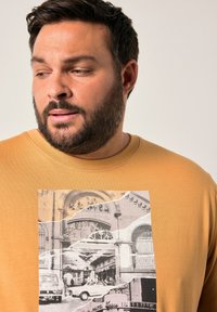 Muž s plnovousem oblečený v hořčicově žluté košili s černobílým vintage tisky městské krajiny, na kterém jsou auta a staré budovy.