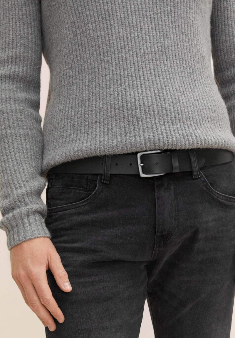 Grau gerippter Pullover mit langen Ärmeln, kombiniert mit schwarzen Jeans und einem schwarzen Ledergürtel mit einer silbernen quadratischen Schnalle.