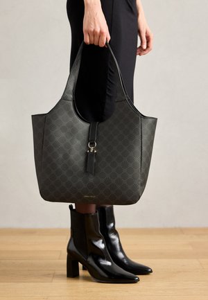 Black patterned tote bag with a smooth texture, slim handles, and a front strap detail. Worn with black ankle boots, set against a wooden floor.