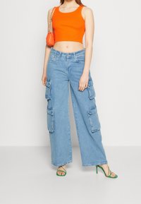 Nainen, jolla on päällään oranssi hihaton crop-top, vaaleansiniset leveät cargo-farkut, vihreät nauhalla kiinnitettävät korot ja pieni oranssi käsilaukku.