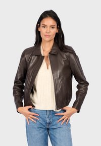 Veste en cuir marron avec un col, fermeture éclair à l'avant et poches latérales. Portée par-dessus un pull en maille léger et un jean bleu.