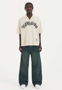 Krátký béžový button-up košile s černým grafickým textem, kombinováno s širokými tmavě zelenými džíny a bílými teniskami.