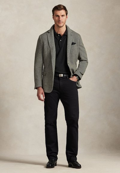 Man standing wearing gray blazer over black polo shirt, black pants, black belt, and black shoes against plain beige background.