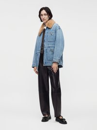 Femme portant une veste en jean bleue avec un col en fourrure beige, un pantalon en cuir noir, une chemise blanche et des mocassins noirs, debout devant un fond uni.