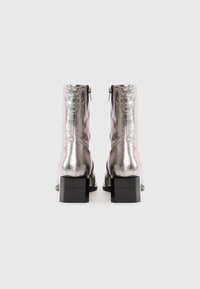 Botines de tobillo plateados metálicos con un acabado brillante, cierre de cremallera y tacón cuadrado. Los acentos incluyen una marca en relieve en el tacón.