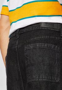 Bakifrån av en person som bär svarta denimjeans med en läderetikett från Urban Classics och en randig skjorta i gult, vitt och teal.