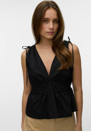 Kobieta o długich brązowych włosach, ubrana w czarny, bezrękawnik z dekoltem w serek, z talią zaakcentowaną marszczeniem i wiązanymi ramiączkami, zestawiony z beżowymi spodniami.