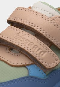 Κοντινή εικόνα των μπεζ δερμάτινων λουριών με βέλκρο σε πολύχρωμο αθλητικό παπούτσι, που αναδεικνύει τις λεπτομερείς ραφές και το ανάγλυφο όνομα της μάρκας.