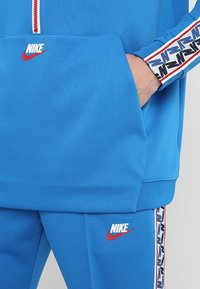 Sudadera azul con capucha y pantalones jogger, ambos con el logotipo de Nike en rojo. Presenta una media cremallera y acentos en negro, blanco y rojo a lo largo de las mangas.