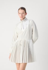 Vit plisserad klänning med hög krage, dragsko i midjan och långa ärmar; har ett strukturerat, slätt tyg och subtil detaljer med logotyp.