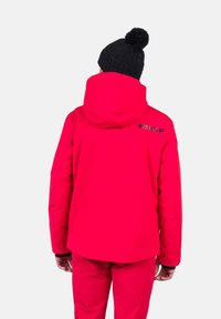 Chaqueta aislante roja con capucha, que cuenta con un sombrero de punto negro texturizado. Logo de "ROSSIGNOL" en impresión oscura en la parte posterior. Tela suave y resistente al agua.