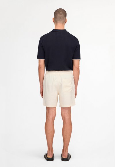 Man standing facing away, wearing a black short-sleeve shirt, beige shorts with back pocket, and black slip-on sandals.