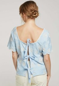 Blouse bleu clair avec des motifs floraux brodés, manches courtes, un dos en scoop profond et deux nœuds pour la fermeture. Le matériau semble texturé.
