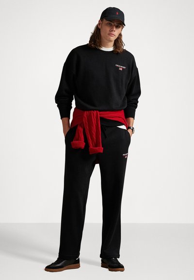 Tânăr îmbrăcat cu un hanorac Polo Sport negru, pantaloni de trening asortați, o șapcă neagră, adidași negri, și un pulover roșu legat în jurul taliei.