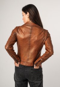 Veste de moto en cuir marron avec un design ajusté, longueur raccourcie, poignets zippés et détails de couture visibles. Portée par-dessus un jean foncé.