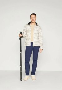 Jasnoszary wzorzysty żakiet z czarnym kołnierzykiem, beżowa bluzka, granatowe spodnie i jasne buty, w zestawie z czarnym kijem trekkingowym.