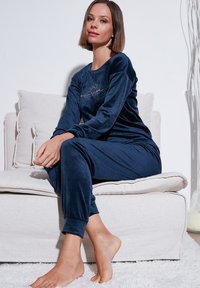 Navy blue velouri pyjama-setti, jossa on pitkät hihat ja hihansuut, ja jota koristaa kultainen brodeerattu aurinkokuvio rinnassa. Henkilö istuu vaalealla sohvall.