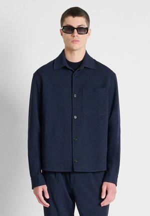 Jonge man met zwarte rechthoekige zonnebril en een donkerblauw overhemd met knopen en bijpassende broek, staand voor een egale lichte achtergrond.