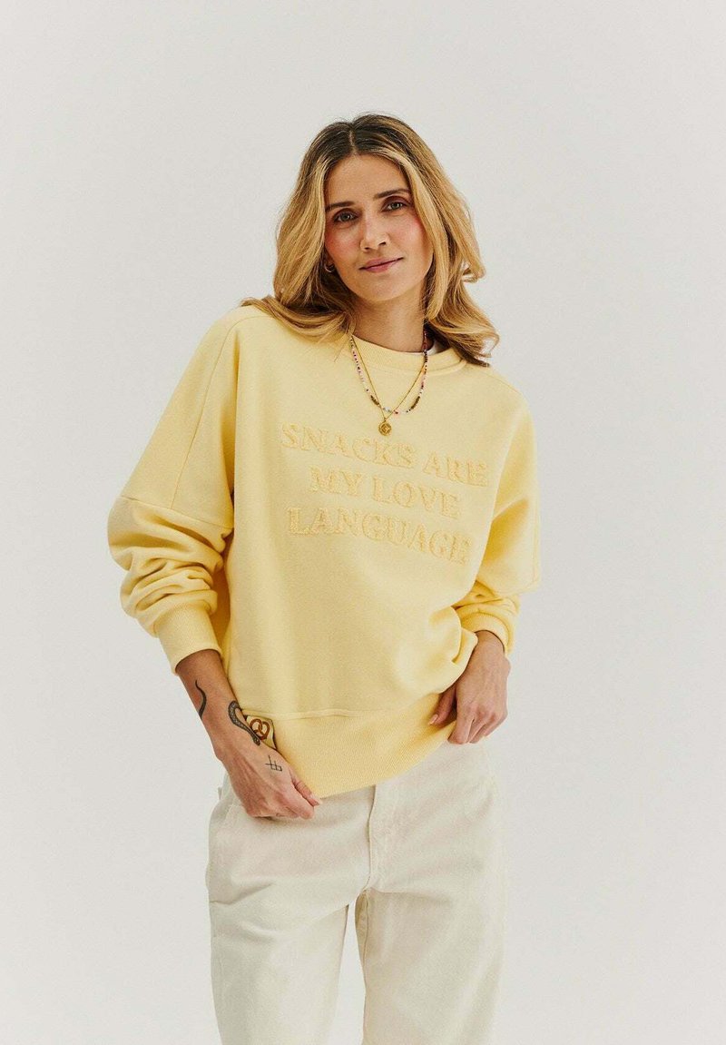 Žltý mikina z fleece s embosovaným textom "SNACKY SÚ MOJÍM JAZYKOM LÁSKY" na prednej strane. Má spadnuté ramenné švy a rebrované manžety.
