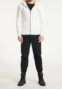 Sweat à capuche zippé blanc avec cordons, poches avant et poignets côtelés, associé à un pantalon cargo noir avec poches latérales et poignets élastiques. Bottes noires.