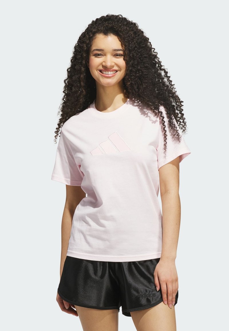 Femme souriante aux longs cheveux bouclés portant un t-shirt Adidas rose clair et un short Adidas noir, debout devant un fond uni.