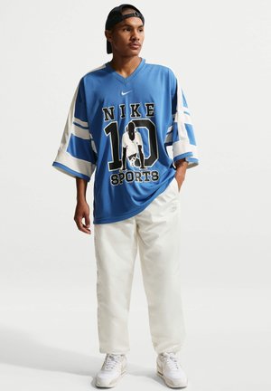 Mann trägt ein blaues übergroßes Nike-Sporttrikot, weiße Hose, weiße Sneakers und eine schwarze rückwärts getragene Kappe, steht mit einer Hand in der Tasche.