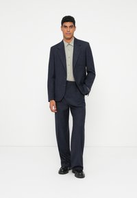 Σκούρο μπλε κοστούμι από υφή ύφασμα, σε συνδυασμό με ανοιχτό πράσινο πουκάμισο με κουμπιά. Το κοστούμι έχει γιακά με εγκοπές και φαρδιά παντελόνια.