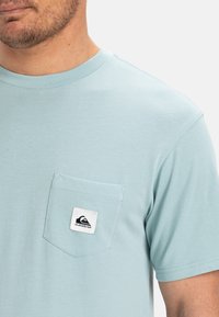 Světle modré tričko z bavlny s kulatým výstřihem a krátkými rukávy. Má náprsní kapsu s černobílou logo nášivkou.