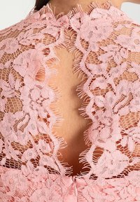 Růžové krajkové šaty s propracovanými květinovými vzory a výstřihem na zádech. Měkká, jemná textura s třepenými okraji a diskrétním zipem.