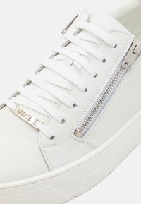 White leather sneaker featuring flat laces, side zipper with metal accents, textured sole, and minimalistic design.