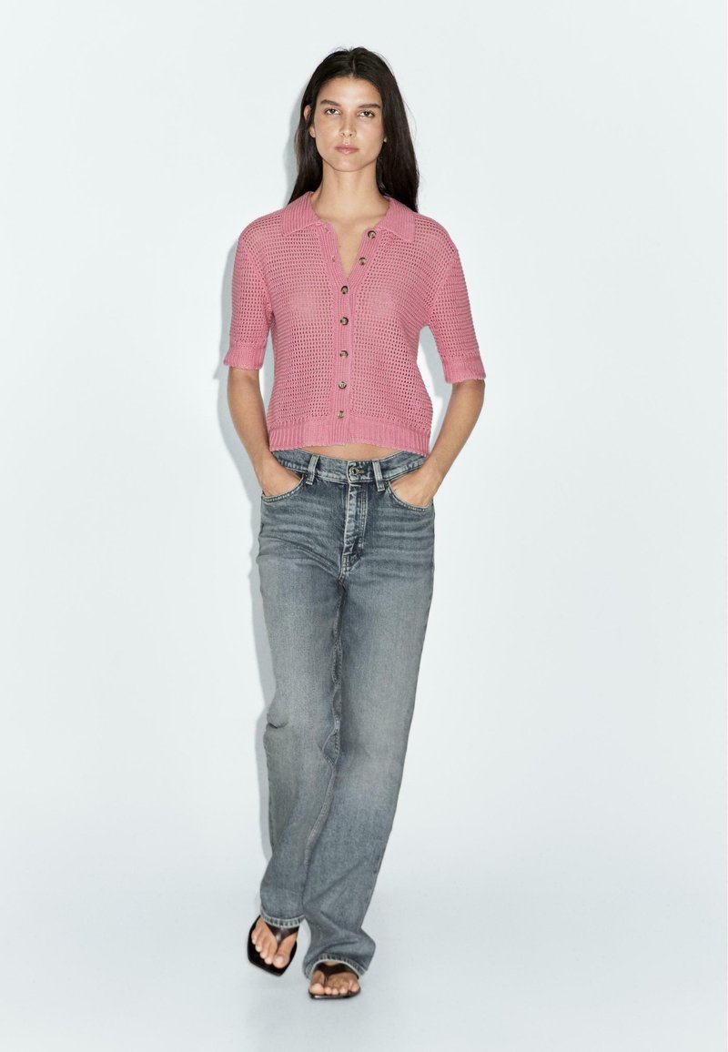 Kurze pinke Strickjacke mit Kragen, Knopfleiste und cropped Länge. Paar hochtaillierte hellblaue Jeans und schwarze Sandalen.