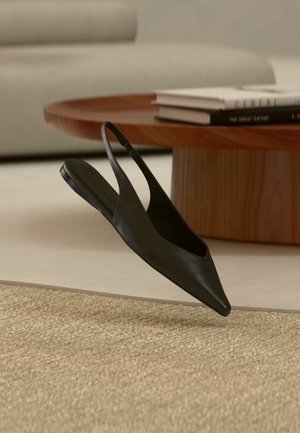 Zwarte platte slingback schoen met puntige neus bij een ronde houten tafel met gestapelde boeken op een beige tapijt in een modern woonkamertje.