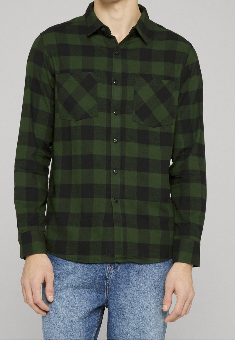 Homme portant une chemise à manches longues à carreaux verts et noirs, boutonnée, avec deux poches poitrine, et un jean bleu sur un fond uni.