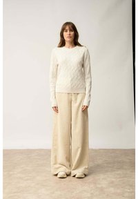 Biała sweter o warkoczowym splocie z długimi rękawami, zestawiony z szerokimi, teksturowanymi beżowymi spodniami. Modelka stoi na neutralnym tle.