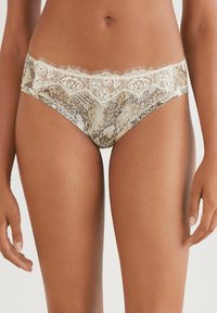 Femme portant des culottes taille basse avec un motif serpent et une bordure en dentelle, se tenant avec les bras détendus le long du corps devant un arrière-plan neutre.