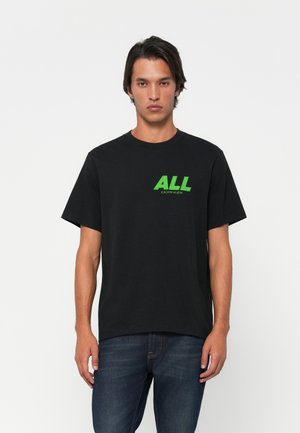Young man wearing a black t-shirt with green "ALL CALVIN KLEIN" text, paired with dark blue jeans, standing against a white background.