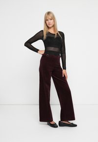 Donna in piedi con una mano sui fianchi, che indossa una maglia trasparente nera a maniche lunghe, pantaloni larghi bordeaux e ballerine nere, su sfondo bianco.