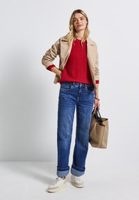 Beige Jacke über einem roten Kabelstrickpullover, blaue hochgekrempelte Jeans und weiße Sneakers. Hält eine beige Tragetasche. Minimalistisches Design.