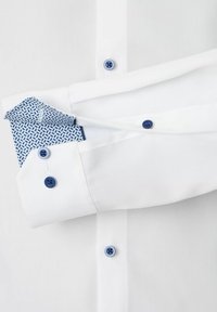 Witte geknopte overhemd met blauwe knopen en blauwe patroon aan de binnenkant van de manchetten, met de mouwen over het borstgebied gevouwen.