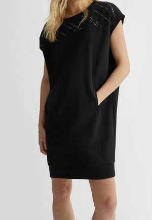 Vestido negro de manga corta con un ajuste relajado, escote redondo y bolsillos laterales; adornado con sutiles detalles metálicos en los hombros.